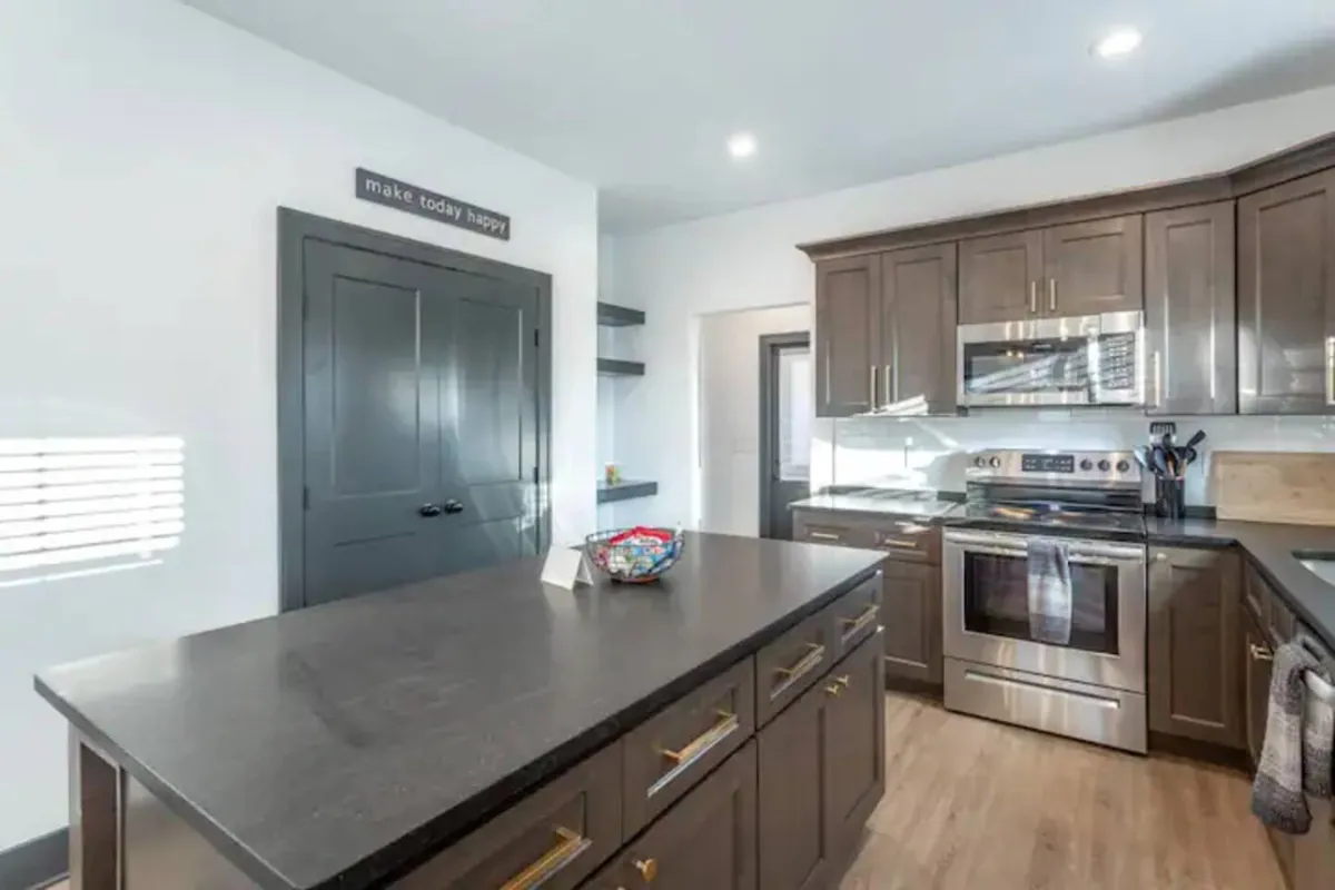 A contemporary kitchen with a central island, stainless steel appliances, dark cabinetry, and motivational wall art. The space feels vibrant and welcoming, designed for meal preparation, entertaining, and family gatherings.