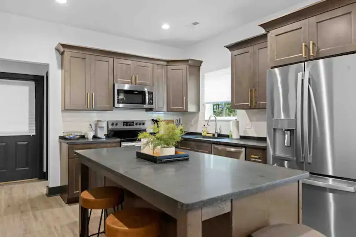 A bright, inviting kitchen interior featuring a prominent kitchen island, sleek stainless steel appliances, dark cabinetry, and a decorative centerpiece on the island. The modern finishes and open layout suggest a space designed for both cooking and socializing, with ample prep areas.