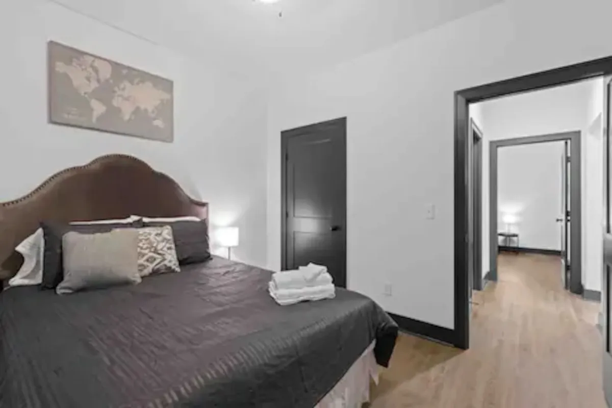 A cozy bedroom with a bed covered in decorative pillows, world map artwork on the wall, and a nightstand with a lamp. The room is brightened by natural light, emphasizing a relaxed and inviting atmosphere.