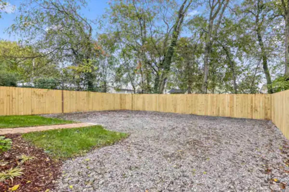A serene backyard space with a wooden fence, gravel surface, and lush greenery. The area is well-suited for outdoor gatherings or letting kids play, with the greenery suggesting a peaceful spot to unwind after a day out.