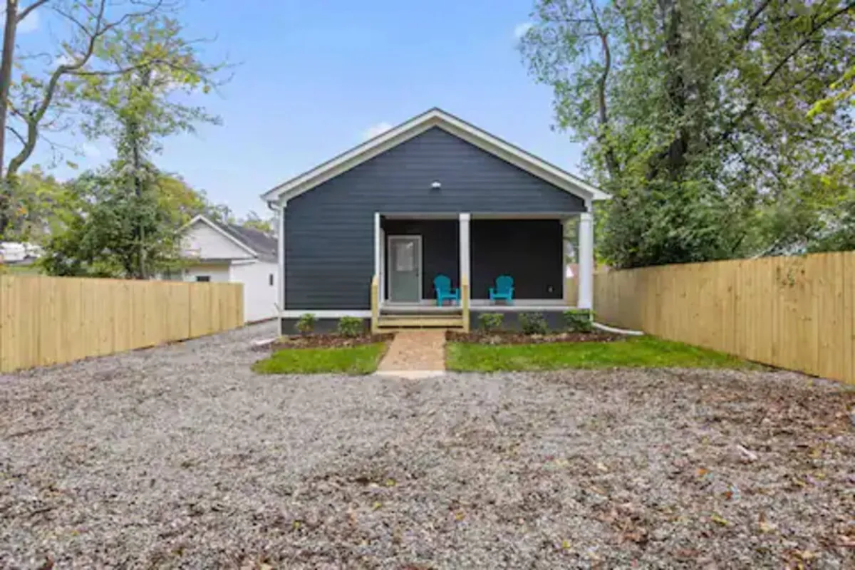 This image features the home's front porch with inviting blue chairs, a gravel driveway, and a wooden fence. The scene is bright and welcoming, suggesting a friendly, secure, and accessible entry to the property. The porch's setup hints at relaxing mornings or evenings, while the well-kept exterior reinforces the new construction and peaceful setting described in the section.
