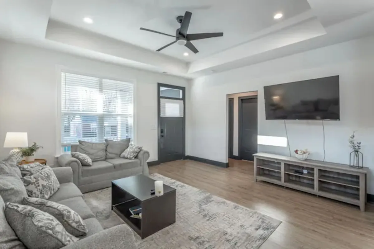 This image features a cozy living area with a sofa, coffee table, television, and the home's front door visible, offering a welcoming view into the heart of the house. Natural light fills the room, and the overall setup invites guests to relax and enjoy time together.