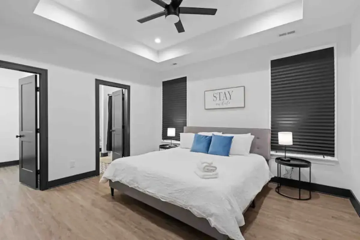 A serene bedroom featuring a king-sized bed adorned with decorative pillows, a 'STAY' wall art piece, and nightstands with lamps on either side. The soft lighting and thoughtful decor create a tranquil atmosphere ideal for unwinding after a busy day.