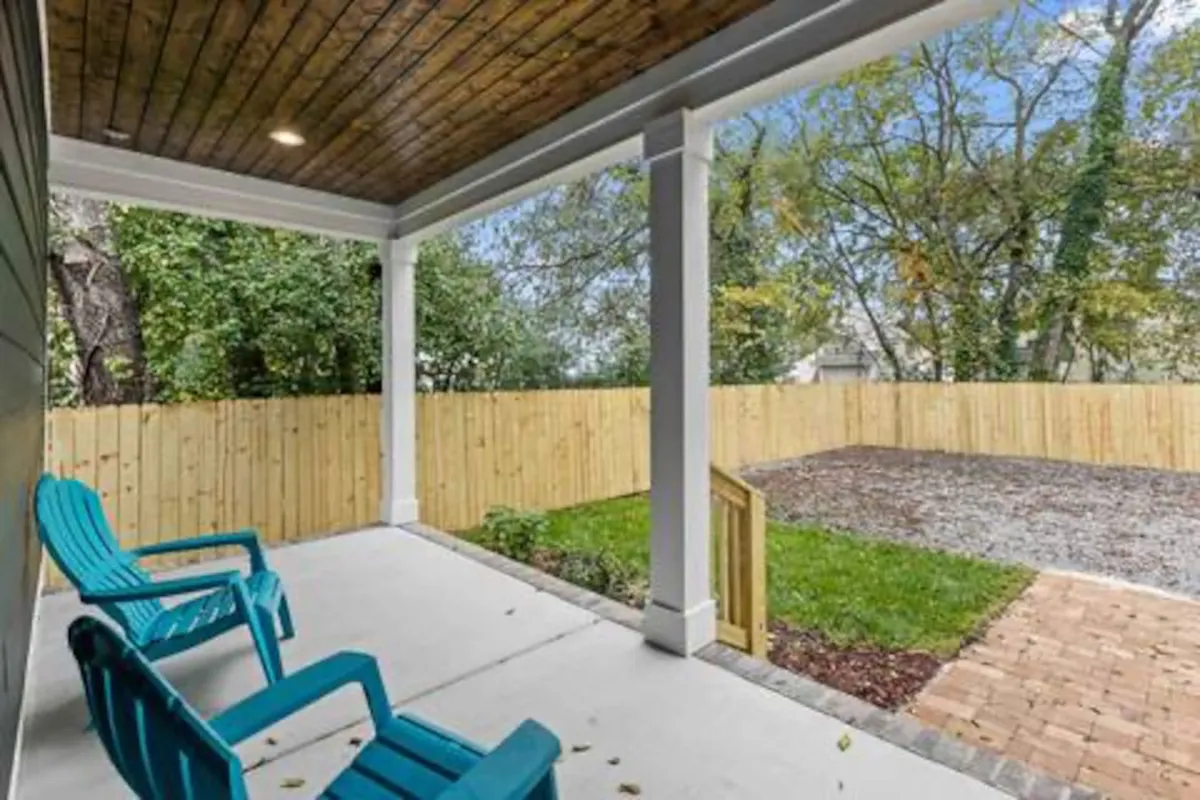 The exterior porch space, highlighted by blue chairs, a wooden ceiling, and lush green vegetation, creates a calm, welcoming atmosphere. The fenced yard offers a safe space for children to play or families to gather before venturing into the city.