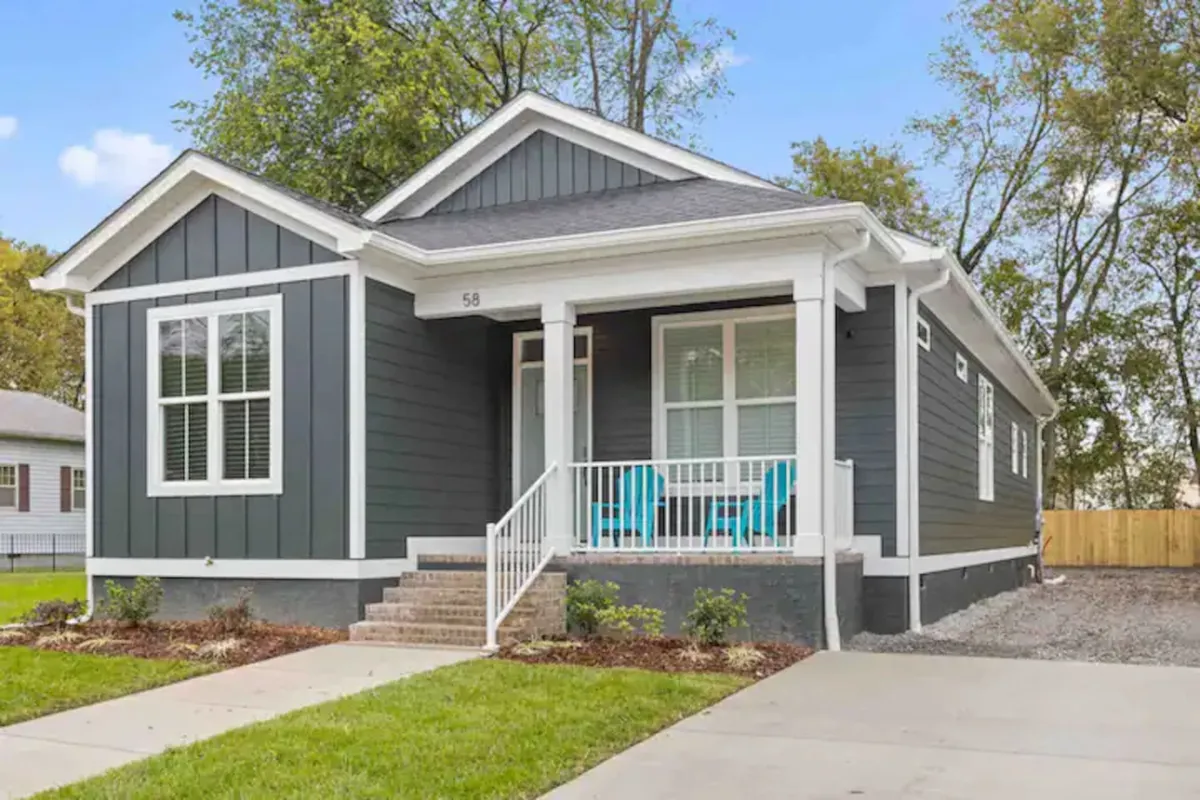The image displays the charming exterior of a new construction single-family house in downtown Chattanooga. The front porch features inviting blue rocking chairs and overlooks a beautifully landscaped yard. Sunlight streams onto the porch, highlighting a welcoming spot for relaxation.